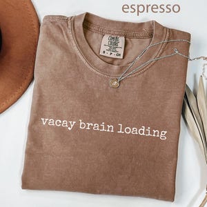 May include: Espresso-coloured t-shirt with the text "vacay brain loading" in white. A silver necklace with a small pendant rests on the shirt. The shirt is folded, and a brown hat is partially visible.