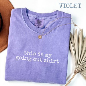 Pode incluir: Camiseta Comfort Colors violeta com o texto branco "this is my going out shirt". Um colar de ouro com um pingente e um chap&eacute;u marrom tamb&eacute;m est&atilde;o na imagem.