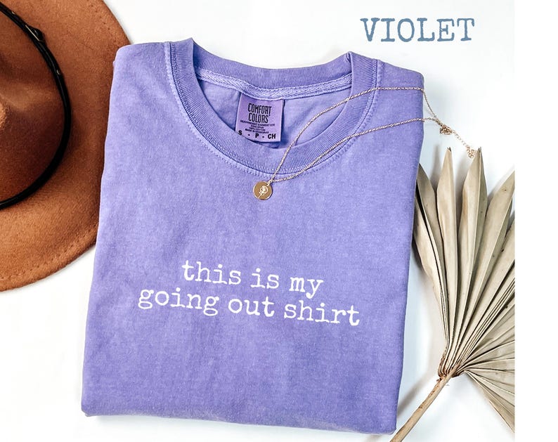 Pode incluir: Camiseta Comfort Colors violeta com o texto branco "this is my going out shirt". Um colar de ouro com um pingente e um chap&eacute;u marrom tamb&eacute;m est&atilde;o na imagem.