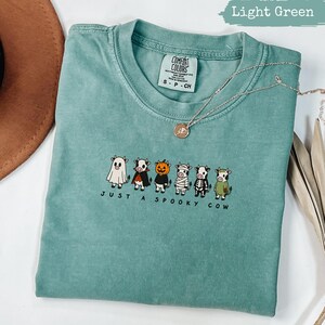 Light green t-shirt with six cows in Halloween costumes and "Just a Spooky Cow" text.
