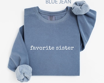 May include: A blue jean-coloured sweatshirt with the words "favorite sister" printed in white. The sweatshirt has a crew neck and long sleeves with folded cuffs. The text "BLUE JEAN" is at the top of the image.