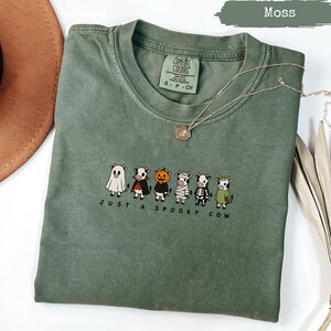 Green t-shirt with "Just a Spooky Cow" text and six cows in Halloween costumes.