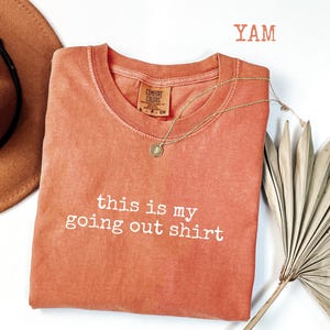 Pode incluir: Uma camiseta Comfort Colors cor coral com a frase "this is my going out shirt" em letras brancas. Um colar de ouro com um pingente est&aacute; drapeado na camiseta. Um chap&eacute;u marrom e uma folha de palmeira seca est&atilde;o no fundo.