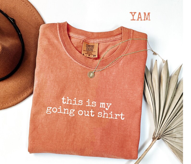 Pode incluir: Uma camiseta Comfort Colors cor coral com a frase "this is my going out shirt" em letras brancas. Um colar de ouro com um pingente est&aacute; drapeado na camiseta. Um chap&eacute;u marrom e uma folha de palmeira seca est&atilde;o no fundo.