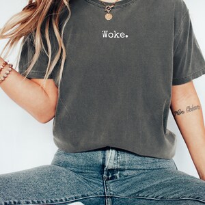 May include: Dark gray t-shirt with the word "Woke" printed in white. The shirt is made of a soft material and has a relaxed fit. The model is wearing blue jeans and a gold necklace.