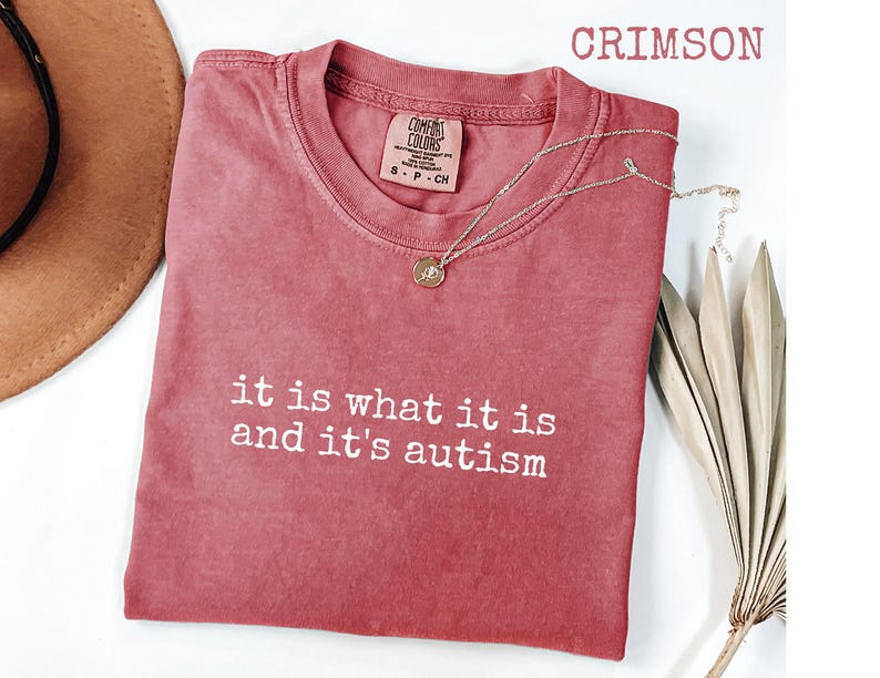 Pu&ograve; includere: T-shirt color cremisi con la scritta bianca "it is what it is and it's autism". Una collana d'oro con un ciondolo &egrave; drappeggiata sulla maglietta. La maglietta &egrave; del marchio Comfort Colors.