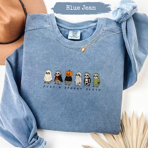 Könnte beinhalten: Ein blaues, jeansfarbenes Sweatshirt mit Rundhalsausschnitt und einer Stickerei von sechs Faultieren in verschiedenen Halloween-Kostümen, darunter ein Geist, Vampir, Kürbis, Mumie, Skelett und Zombie, mit dem Text Just A Spooky Sloth.