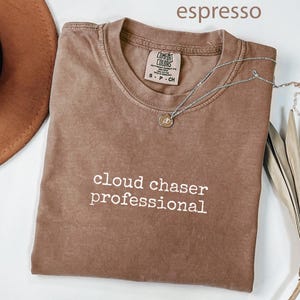 May include: Espresso-colored t-shirt with the words "cloud chaser professional" in white. A silver necklace with a small pendant rests on the shirt. A brown hat and dried plant are also in the image.