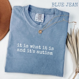 Pu&ograve; includere: T-shirt blu jeans con la scritta bianca "it is what it is and it's autism". La maglietta ha girocollo e maniche corte. Una collana d'oro con un ciondolo &egrave; drappeggiata sulla maglietta.