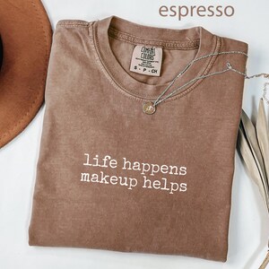 Pode incluir: T-shirt cor de café com o texto branco "life happens makeup helps". Um colar de prata com um pequeno pingente repousa sobre a camisa. A camisa está dobrada e um chapéu marrom é visível no fundo.
