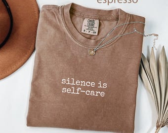 May include: Espresso-colored t-shirt with the text "silence is self-care" in white. The shirt has a crew neck and a small tag on the collar. A silver necklace with a small pendant is draped across the shirt.