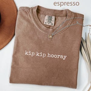 May include: An espresso-colored short-sleeve t-shirt with a crew neckline. The shirt features the white text 'kip kip hooray' printed across the chest. A Comfort Colors brand label is visible at the inner neck, indicating a size small.