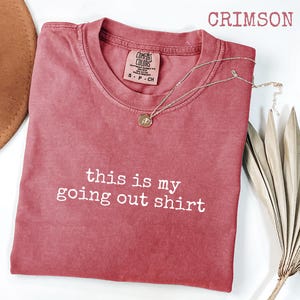 Pode incluir: Camiseta cor carmesim com o texto branco "this is my going out shirt". Um colar dourado com um pingente est&aacute; sobre a camiseta. Um chap&eacute;u marrom e uma planta seca est&atilde;o no fundo.