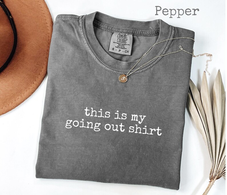 Pode incluir: Uma camiseta cinza Comfort Colors com o texto "this is my going out shirt" em branco. Um colar de prata com um pingente est&aacute; na camisa. Um chap&eacute;u marrom e folhas de palmeira secas est&atilde;o no fundo.