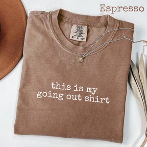 Pode incluir: Camiseta cor espresso com o texto branco "this is my going out shirt". Um colar de prata com um pequeno pingente repousa sobre a camiseta. A camiseta &eacute; da marca Comfort Colors.