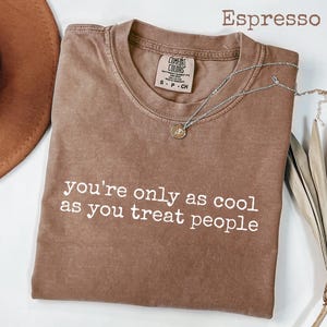 Puede incluir: Camiseta color espresso con el texto blanco "you're only as cool as you treat people". Un collar plateado con un pequeño colgante descansa sobre la camiseta doblada. Un sombrero marrón es parcialmente visible.