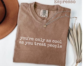 Op de afbeelding: Espresso-kleurig T-shirt met de witte tekst "you're only as cool as you treat people". Een zilveren ketting met een kleine hanger ligt op het gevouwen shirt. Een bruine hoed is gedeeltelijk zichtbaar.