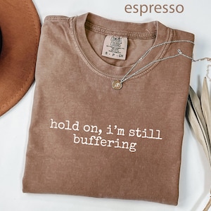 May include: Espresso-coloured t-shirt with the text "hold on, i'm still buffering" in white. A silver necklace with a small pendant rests on the shirt. A brown hat and dried plant are in the background.