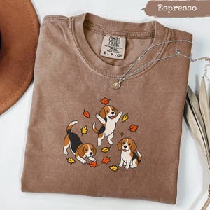 Puede incluir: Una camiseta color espresso con un gráfico de tres perros beagle jugando con hojas de otoño dispersas. La camiseta tiene cuello redondo y una etiqueta en el interior del cuello que dice Comfort Colors. Una cadena plateada descansa sobre la tela.