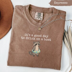 Pode incluir: Camiseta cor espresso com a frase "it's a good day to drink on a boat" e um guaxinim de desenho animado usando uma boia floral. A camiseta tem gola redonda e mangas curtas. Um colar de prata está drapeado sobre a camiseta.