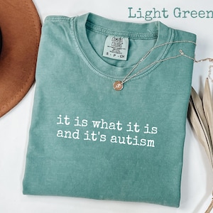 Pu&ograve; includere: T-shirt verde chiaro con la scritta "it is what it is and it's autism" in bianco. Una collana d'oro con un ciondolo &egrave; appoggiata sulla maglietta. La maglietta &egrave; piegata e un cappello marrone &egrave; parzialmente visibile.