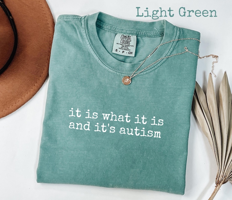 Pu&ograve; includere: T-shirt verde chiaro con la scritta "it is what it is and it's autism" in bianco. Una collana d'oro con un ciondolo &egrave; appoggiata sulla maglietta. La maglietta &egrave; piegata e un cappello marrone &egrave; parzialmente visibile.