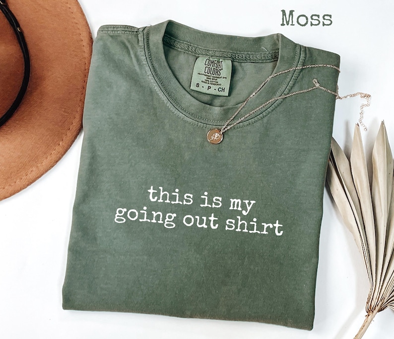 Pode incluir: Uma camiseta verde musgo com o texto "this is my going out shirt" em branco. Um colar dourado com um pequeno pingente repousa sobre a camiseta. Um chap&eacute;u marrom e uma planta seca est&atilde;o no fundo.