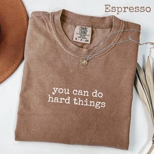 May include: Espresso-coloured t-shirt with the text "you can do hard things" in white. A silver necklace with a small pendant rests on the shirt. The shirt is a Comfort Colors brand.