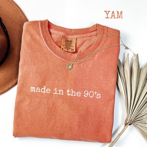 Puede incluir: Una camiseta de color coral con la frase "made in the 90's" bordada en blanco. Un collar dorado con un colgante est&aacute; drapeado sobre la camiseta. Un sombrero marr&oacute;n y una hoja de palma seca tambi&eacute;n est&aacute;n en la imagen.