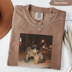 Può includere: T-shirt Comfort Colors color Espresso con una grafica di una tartaruga con occhiali da sole che beve caffè freddo. La maglietta ha girocollo e maniche corte. Una collana d'argento con un piccolo ciondolo è drappeggiata sulla maglietta.