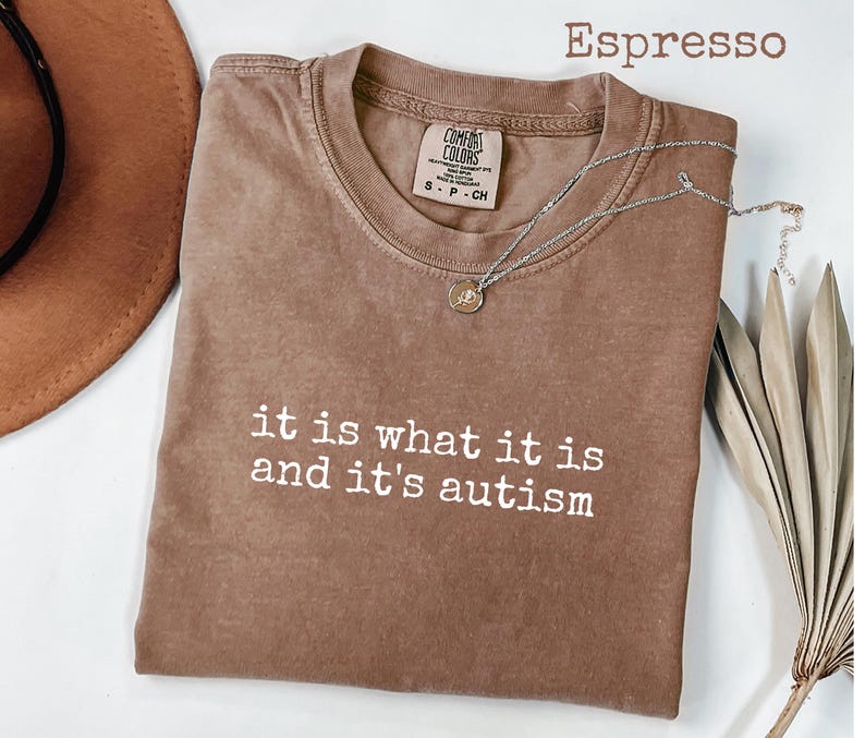 Pu&ograve; includere: T-shirt color espresso con la scritta bianca "it is what it is and it's autism". Una collana d'argento con un piccolo ciondolo &egrave; drappeggiata sulla maglietta. La maglietta &egrave; piegata e l'etichetta Comfort Colors &egrave; visibile.