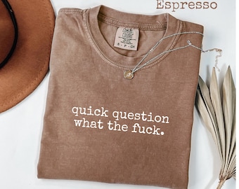 Può includere: T-shirt color espresso con la scritta "quick question what the fuck." Una collana d'argento con un piccolo ciondolo è appoggiata sulla maglietta. La maglietta è del marchio Comfort Colors.