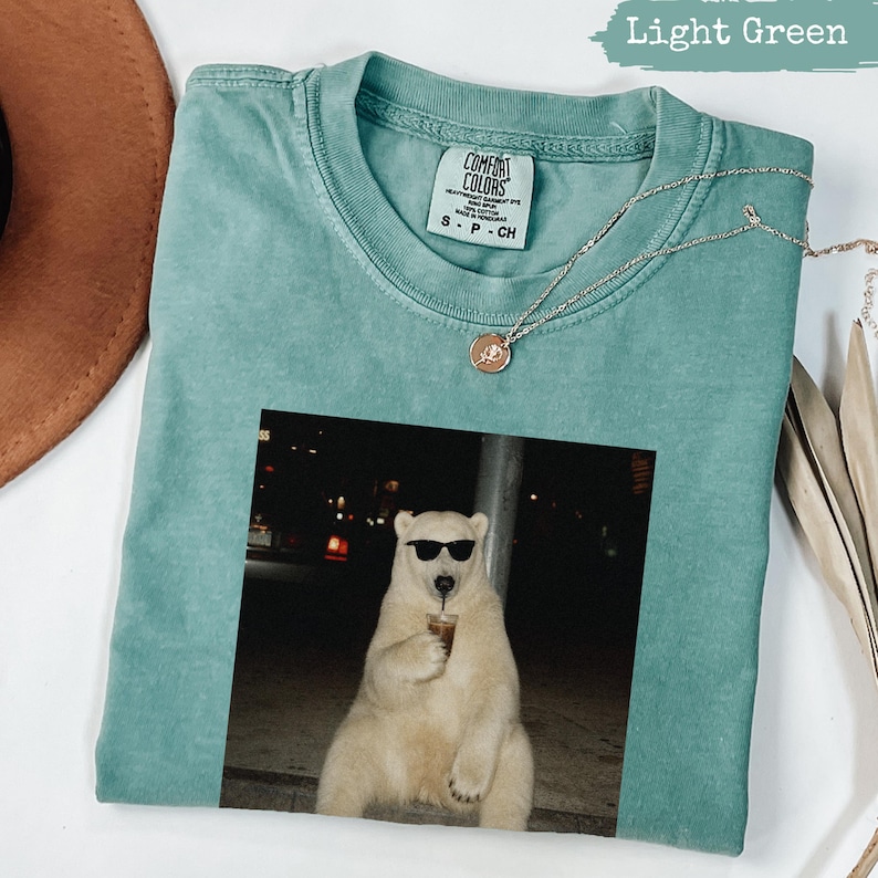 Light green t-shirt with a graphic of a bear wearing sunglasses holding a drink.