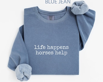 May include: A blue jean-coloured sweatshirt with the white text "life happens horses help". The sweatshirt has a crew neck and long sleeves with a rolled cuff. The top is made of a soft material.