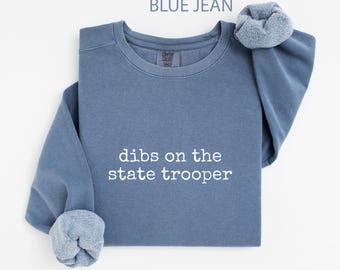 May include: A blue jean-coloured sweatshirt with the white text "dibs on the state trooper". The sweatshirt features a crew neck and ribbed cuffs. The sleeves are rolled up, revealing a lighter blue lining.