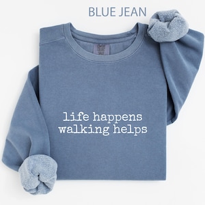 May include: A blue jean coloured sweatshirt with the text "life happens walking helps" in white. The sweatshirt has a crew neck and the sleeves are cuffed. The Comfort Colors brand tag is visible.
