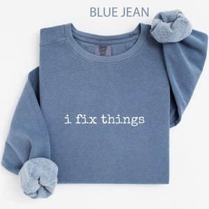 May include: Blue jean-colored sweatshirt with the text "i fix things" in white. The sweatshirt has a crew neck and long sleeves with rolled-up cuffs. The Comfort Colors brand tag is visible on the upper back.