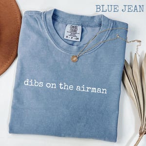 May include: A blue jean colored t-shirt with the text "dibs on the airman" in white. The shirt has a crew neck and short sleeves. A gold necklace with a pendant is draped on the shirt. The shirt is folded on a white surface.