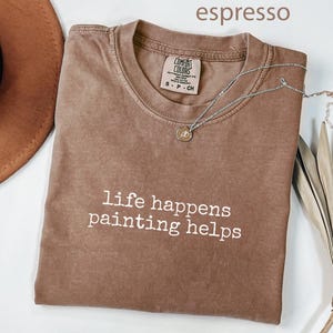 Könnte beinhalten: Espressofarbenes T-Shirt mit dem weißen Text "life happens painting helps". Eine silberne Halskette mit einem kleinen Anhänger liegt auf dem Shirt. Das Shirt ist gefaltet, und ein brauner Hut ist teilweise sichtbar.