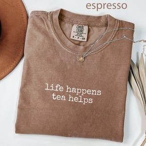 May include: Espresso-colored t-shirt with the text "life happens tea helps" in white. A silver necklace with a small pendant rests on the shirt. The shirt is folded, and a brown hat is partially visible.