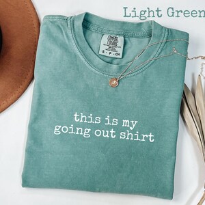 Pode incluir: Camiseta verde claro com o texto branco "this is my going out shirt". Um colar dourado com um pingente repousa sobre a camiseta. Um chap&eacute;u marrom e uma planta seca est&atilde;o no fundo.