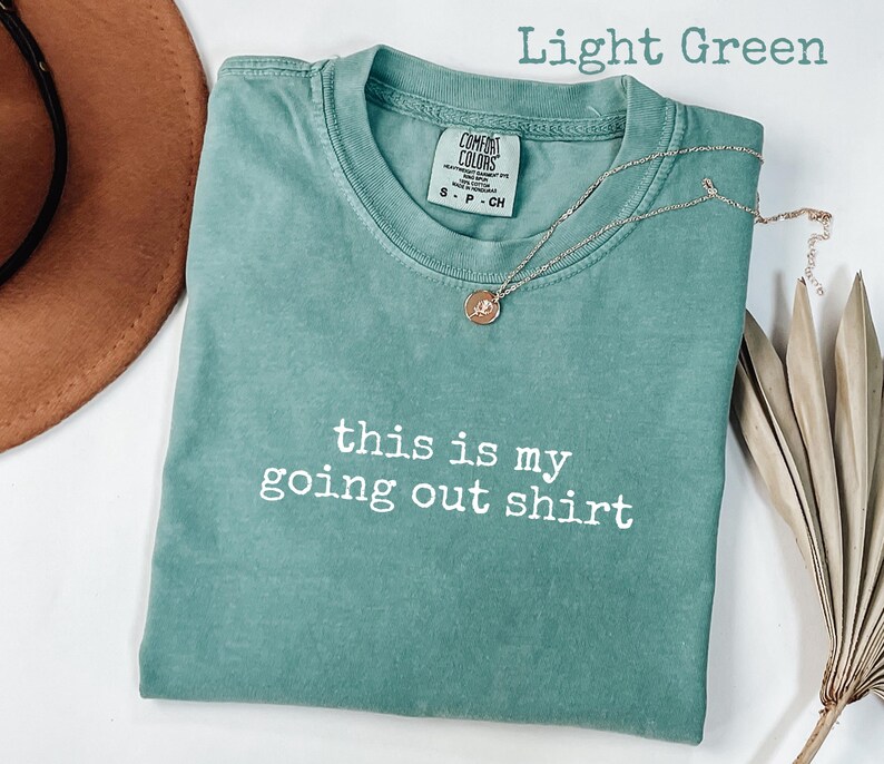 Pode incluir: Camiseta verde claro com o texto branco "this is my going out shirt". Um colar dourado com um pingente repousa sobre a camiseta. Um chap&eacute;u marrom e uma planta seca est&atilde;o no fundo.