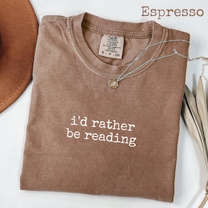 Puede incluir: Camiseta color espresso con la frase blanca "i'd rather be reading". Un collar plateado con un pequeño colgante descansa sobre la camiseta. La camiseta es de la marca Comfort Colors.