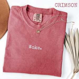 May include: Crimson-colored t-shirt with the word "Woke" embroidered in white. A delicate silver necklace with a pendant rests on the shirt. The shirt is from the Comfort Colors brand. A brown hat and dried plant are in the background.