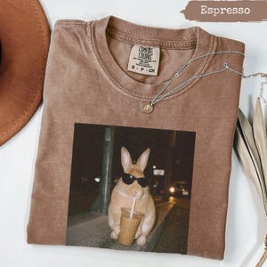 May include: Espresso-colored Comfort Colors t-shirt with a graphic of a rabbit wearing sunglasses and drinking from a cup. The shirt has a crew neck and short sleeves. A silver necklace with a pendant is draped across the shirt.