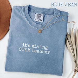 Puede incluir: Camiseta azul vaquero con el texto "it's giving STEM teacher" en blanco. La camiseta tiene cuello redondo y mangas cortas. Un collar dorado con un colgante está drapeado sobre la camiseta. La camiseta está hecha de un material suave y cómodo.