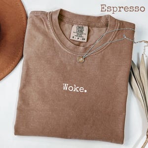 May include: Espresso-colored t-shirt with the word "Woke" embroidered in white. The shirt has a crew neck and short sleeves. A silver necklace with a small pendant is draped across the shirt.