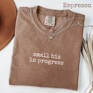 Puede incluir: Camiseta color espresso con el texto "small biz in progress" en blanco. Un collar plateado con un pequeño colgante descansa sobre la camiseta. La camiseta es de la marca Comfort Colors.