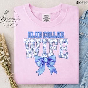May include: A light pink t-shirt with the words "BLUE COLLAR WIFE" in blue block letters with a floral design. A blue bow is below the text. The shirt is from the brand Comfort Colors.