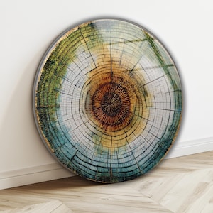 May include: Round wall art featuring a detailed cross-section of a tree trunk. The artwork displays concentric rings in shades of brown, yellow, green, and blue, with a dark center. The piece has a metallic gold edge.